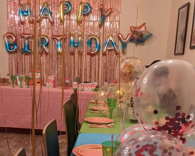 A dining table is set for a birthday celebration, featuring colorful plates, glasses, and decorated balloons, with a "HAPPY BIRTHDAY" banner displayed in the background.