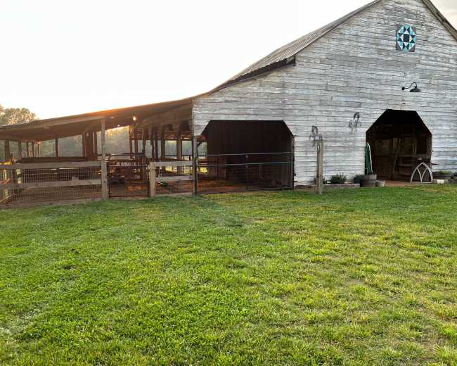 A weathered barn sits on a grassy field, with sunlight illuminating one side and an adjoining fenced area.