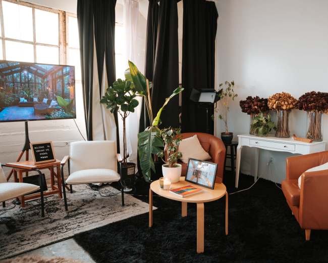 The image shows a cozy interior space featuring two brown armchairs, a round coffee table, a television, various indoor plants, and a side table adorned with dried flowers.