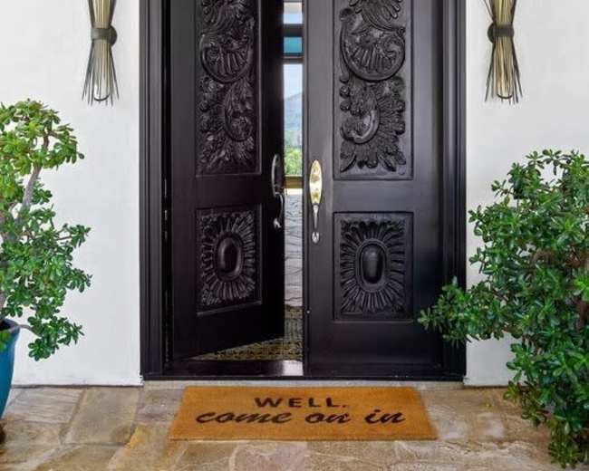 A stylish black double door with intricate carvings stands open, revealing an entranceway adorned with a welcome mat.