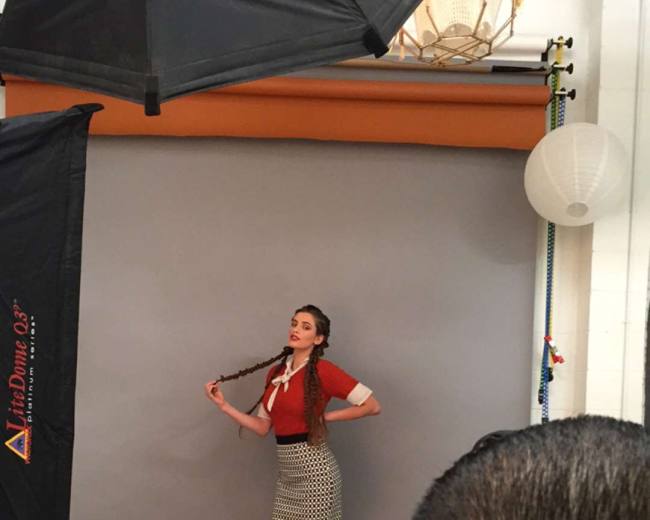 A model poses in front of a gray backdrop while a photographer captures the shot from behind.