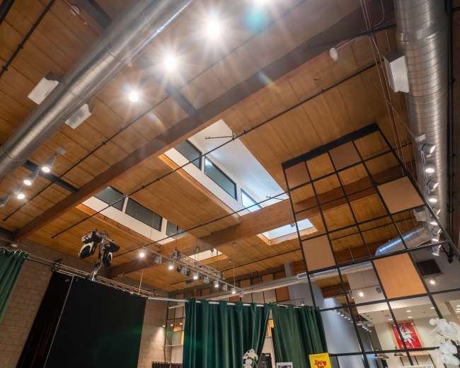 The image shows a modern interior space with exposed wooden beams, track lighting, and large green curtains, featuring a balcony area above.