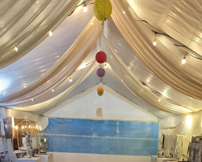 The image shows a tented space with draped fabric overhead, wooden flooring, and several tables lined along the sides, illuminated by string lights and colorful hanging lanterns.