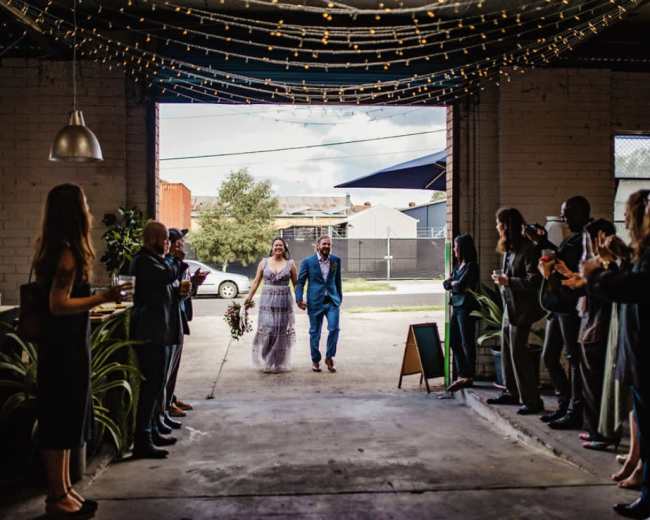 A bride and groom walk hand in hand through a doorway, greeted by guests clapping on either side in an industrial venue.