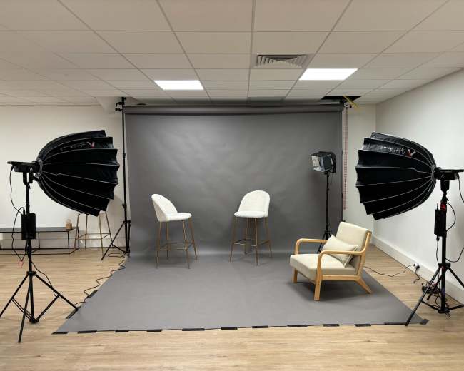 The image shows a photography studio setup with two high stools, a chair, and studio lighting on either side of a gray backdrop.