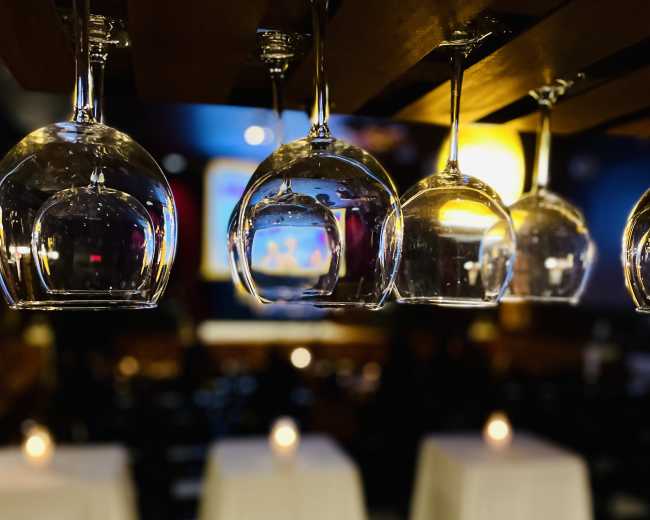 The image shows a dining area with tables covered in white tablecloths and wine glasses hanging upside down from a wooden rack above.