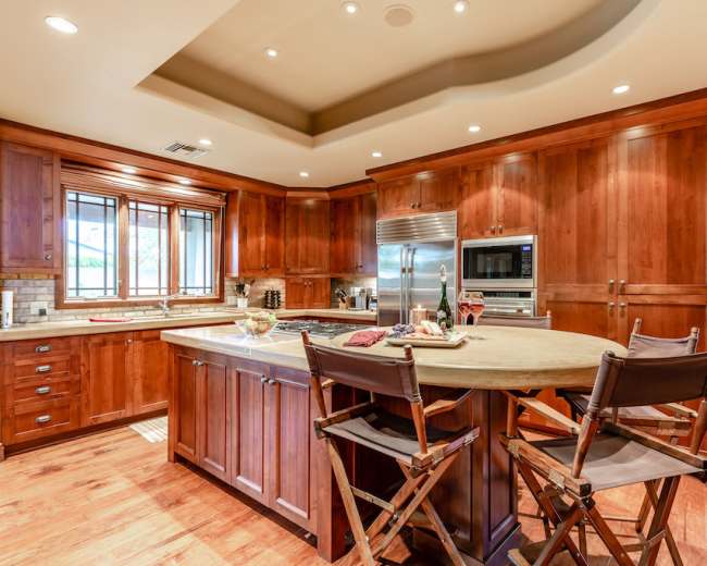 The image shows a modern kitchen with wooden cabinets, a large island with seating, and stainless steel appliances.
