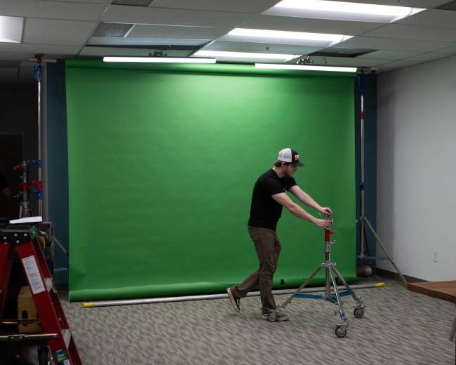 A person adjusts a tripod in front of a green backdrop in a room with carpeted flooring.
