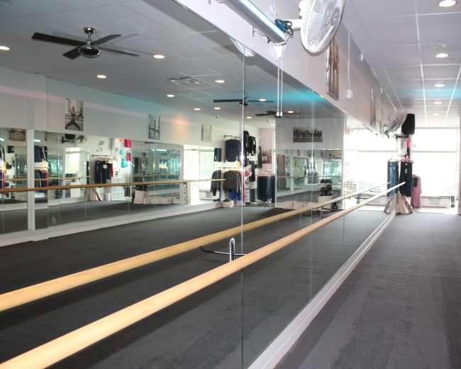 A spacious dance studio features mirrored walls, a wooden ballet bar, and a black carpeted floor.