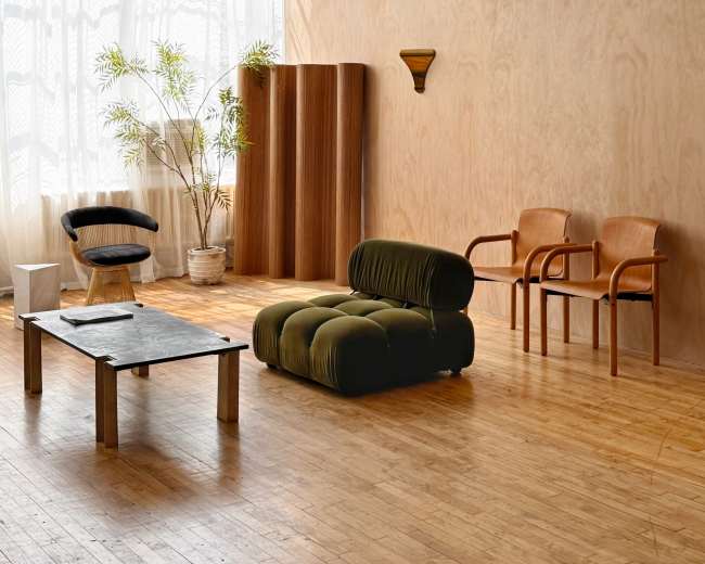 The image shows a minimalist room with wooden walls, a low velvet green sofa, a round black coffee table, two wooden chairs, and a potted plant near large windows.