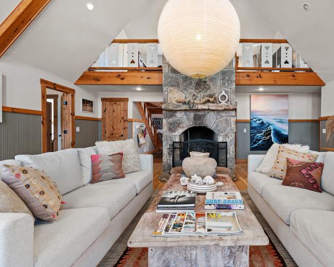 A spacious living room features two light-colored sofas facing a stone fireplace, with a large round pendant light overhead and a coffee table adorned with magazines and decorative items.