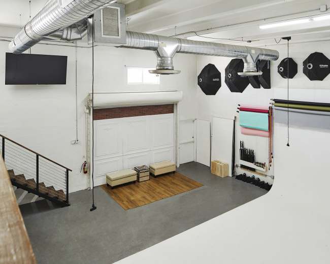 The image shows a spacious photography studio featuring a white backdrop, various colored backdrops, equipment stands, and a seating area.