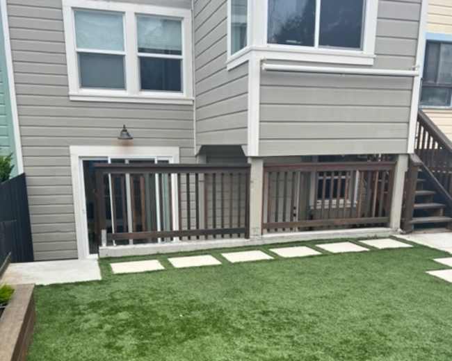 The image shows a backyard with artificial grass, concrete stepping stones, and a wooden railing adjacent to a house with multiple windows.