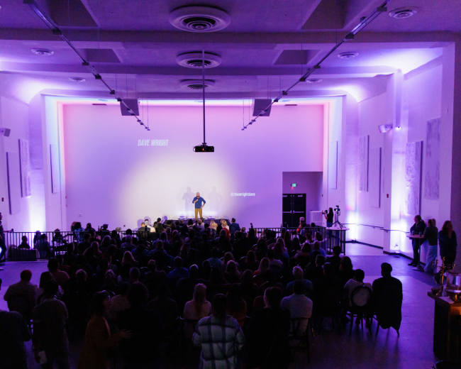 A speaker stands on stage in a well-lit auditorium filled with an audience, with a large screen displaying text in the background.