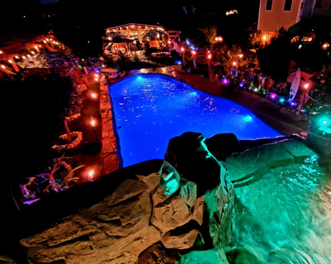A lit outdoor pool area with colorful lights, a waterfall feature, and a gathering space in the background.