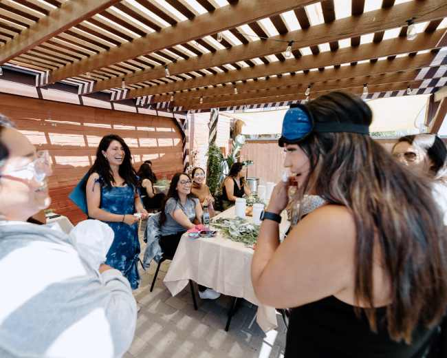 A group of women gathers under a wooden pergola with tables set for an event.