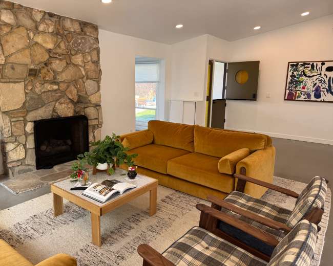 The image shows a cozy living room with a stone fireplace, a yellow sofa, two plaid armchairs, and a wooden coffee table on a light rug.