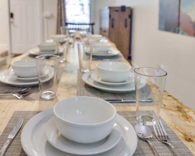 A long dining table is set with white dishes, glasses, and utensils, ready for a meal.