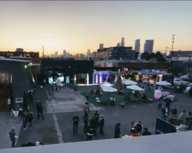 The image shows an outdoor event space at dusk with people gathered, food stalls, and city skyline in the background.