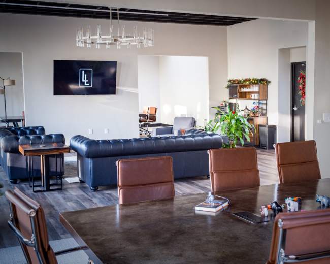 The image shows a modern open-concept office space with a large sectional sofa, a dining table, and a wall-mounted screen displaying a logo.