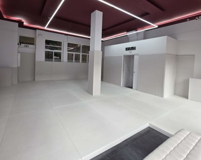 The image depicts a modern, minimalistic interior space with white padded walls, a dark tiled floor, and a ceiling featuring geometric lighting.