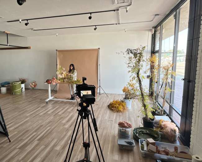 A photographer is set up in a bright studio with multiple cameras focused on a woman arranging dried flowers against a backdrop.