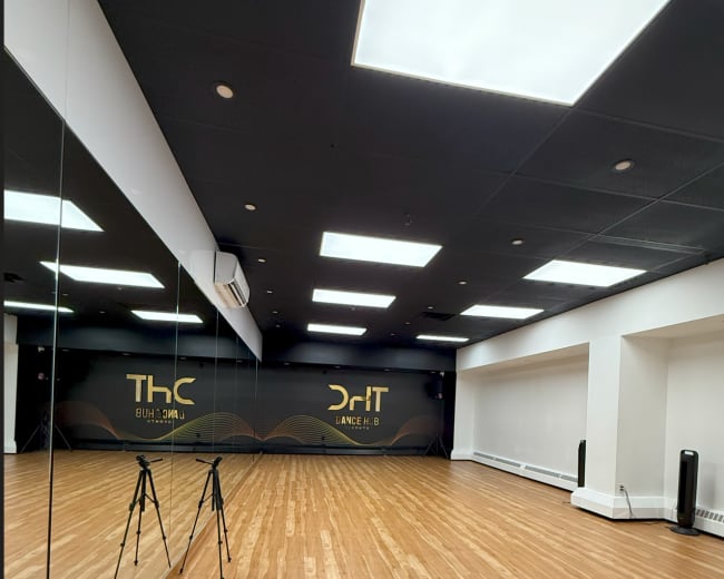 The image shows a spacious dance studio with wooden flooring, bright ceiling lights, and a mirrored wall featuring the names "ThC" and "DrIT" on one side.