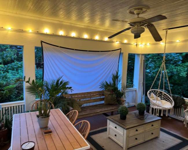 The image shows a covered porch with a wooden table, a hanging chair, and decorative string lights illuminating the space.