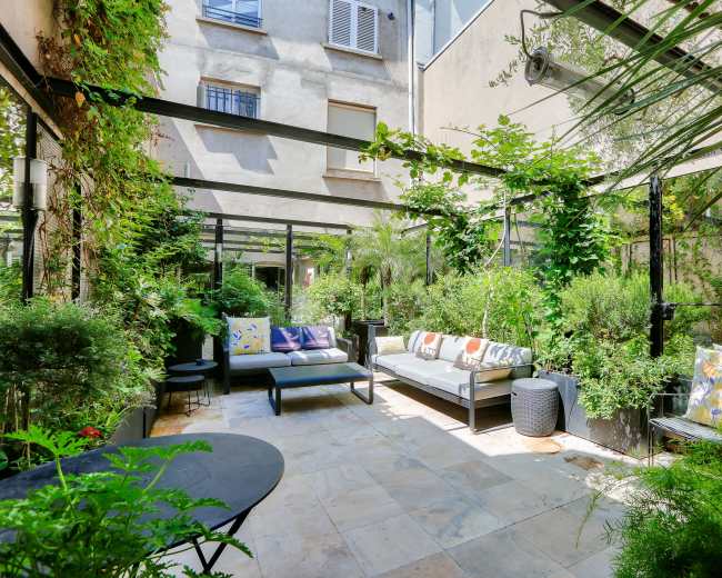 A shaded courtyard features a mix of potted plants, seating areas, and decorative pillows surrounded by greenery.