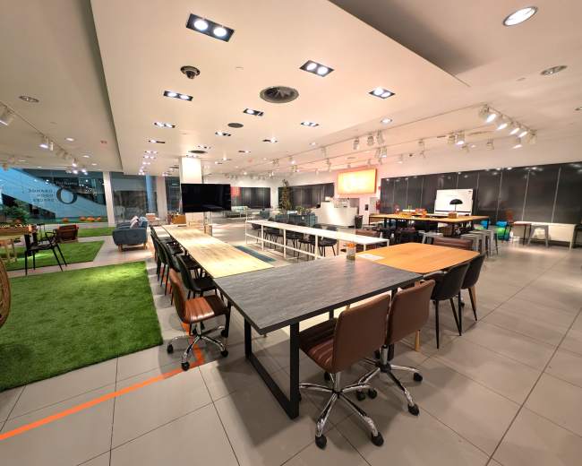 The image shows a modern furniture showroom featuring various tables and chairs, with a mix of seating areas and green decor.