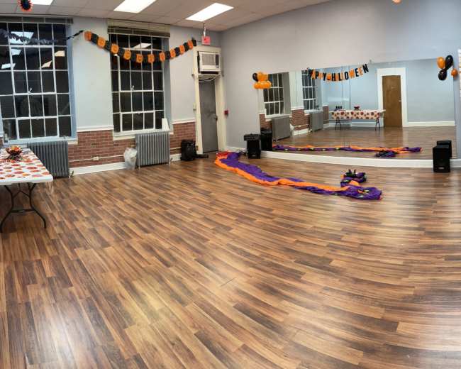 The image shows a spacious indoor room decorated for Halloween with orange and black decorations, a table with festive items, and purple streamers on the floor.