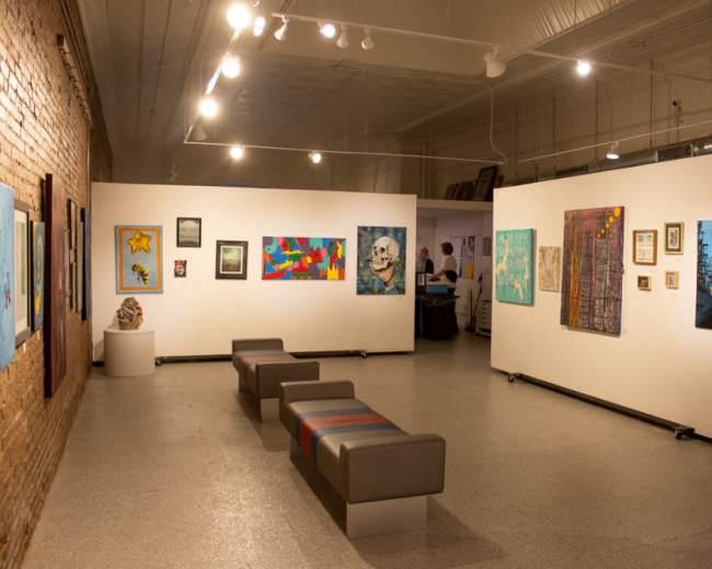 The image shows an art gallery with several colorful artworks displayed on white walls and a few gray benches arranged in the open space.