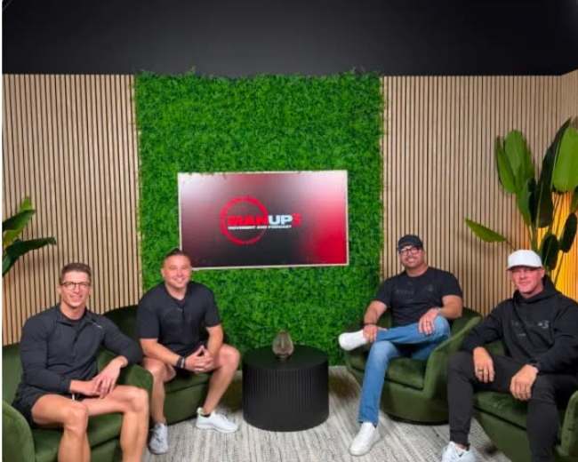 Four men sit on green couches in a studio setting with a large screen displaying "MAN UP" on a green wall adorned with plants.