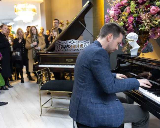 A man plays a grand piano in front of an audience surrounded by flower arrangements and elegant decor.