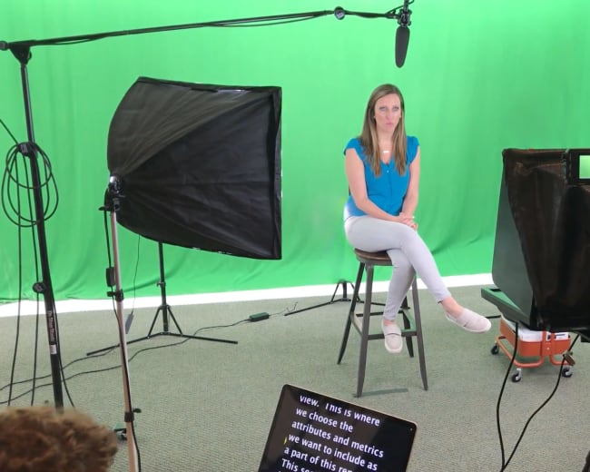 A woman sits on a stool in front of a green screen, surrounded by lighting equipment and a camera setup.