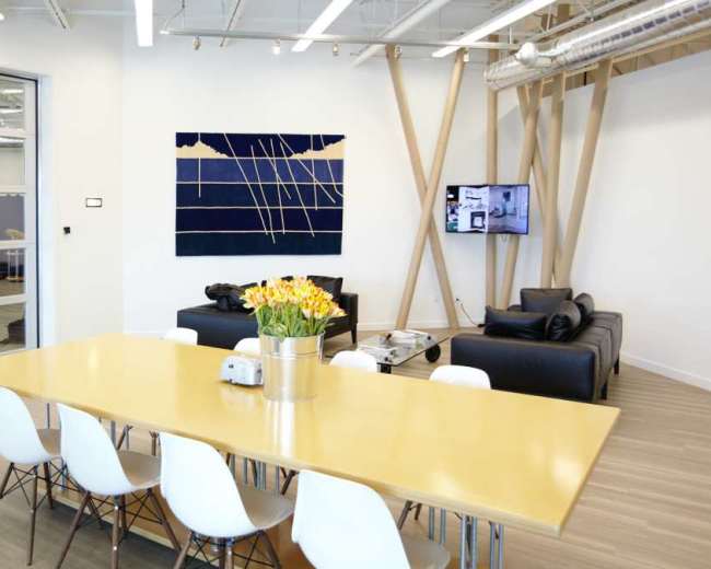 The image shows a modern office space with a long yellow dining table, white chairs, and a seating area featuring black sofas against a backdrop of wood and large windows.