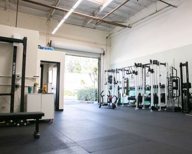 A spacious gym interior with various exercise equipment arranged along the walls and an open garage door leading to an outdoor area.