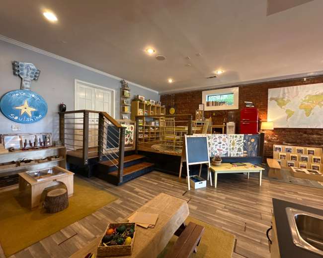 The image shows a brightly lit room with wooden flooring, featuring a play area with shelves stocked with toys, a staircase, a map on the wall, and various learning materials arranged on tables.