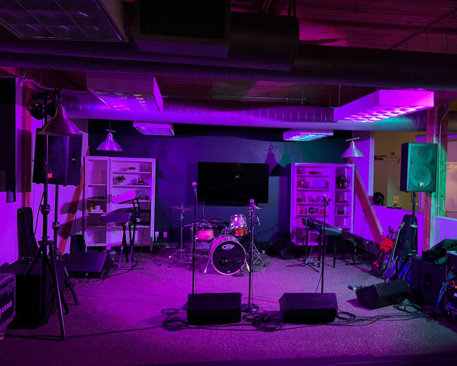 A music stage setup with a drum kit, microphones, and various musical instruments in a venue featuring purple lighting.