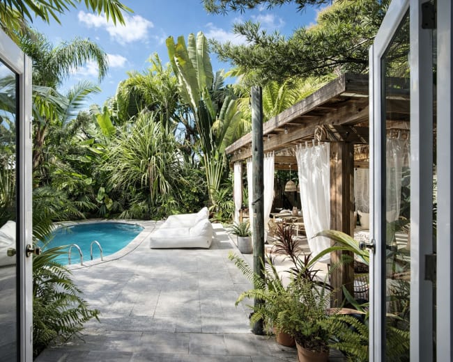 A view from an open doorway reveals a pool surrounded by lush greenery and a wooden structure with seating.