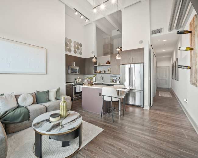 The image shows a modern, open-concept living space with a sofa, round coffee table, and a kitchen featuring stainless steel appliances and pendant lighting.