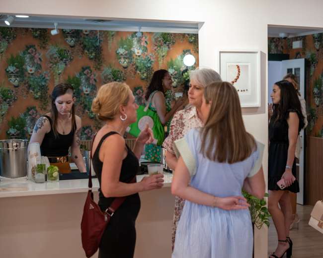A group of women socialize in a brightly decorated room with floral wallpaper, while some are engaged at a counter preparing drinks.