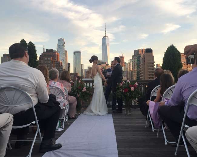 A couple exchanges vows at an outdoor wedding ceremony with a city skyline in the background.