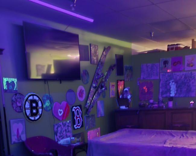 The image shows a colorful, dimly lit room decorated with various artworks and illuminated by purple lighting, featuring a large bed and a set of black chairs with a zebra print table.