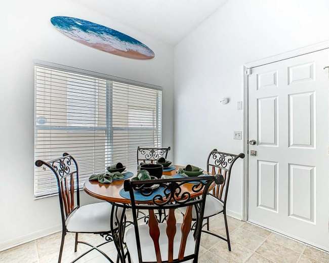 The image shows a small dining area with a round table set for four and a surfboard mounted on the wall above a window.
