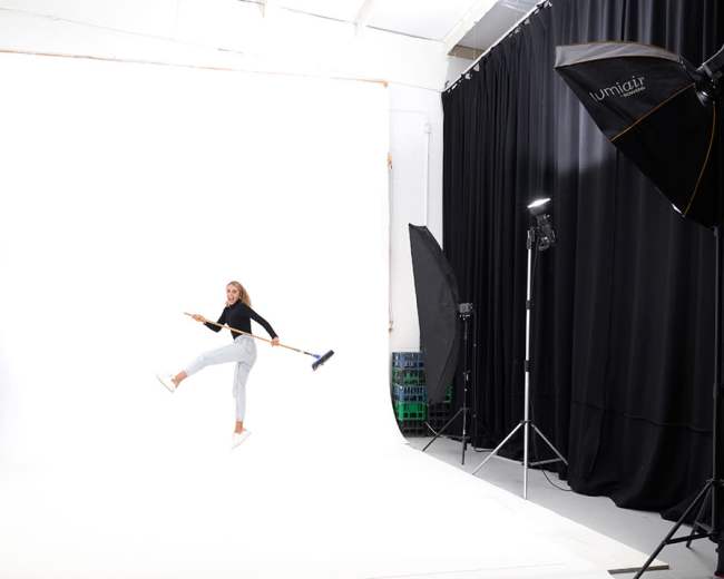 A person is playfully holding a broom while jumping in a photography studio with a white backdrop and lighting equipment.