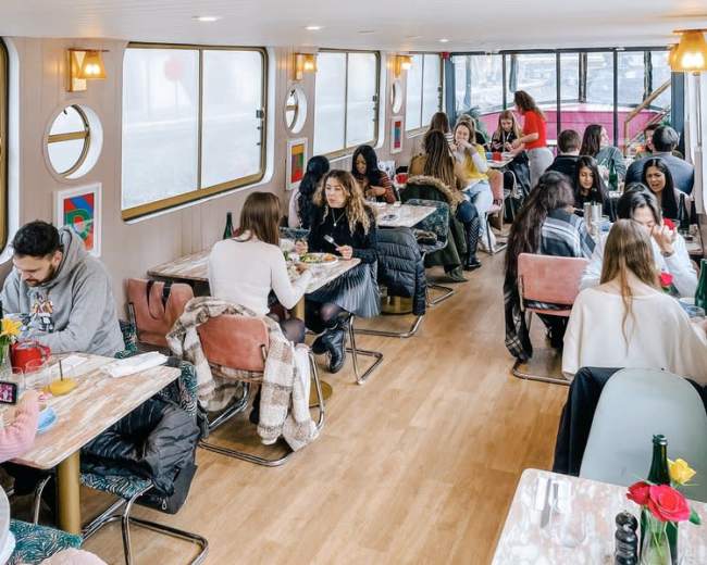 The image shows a bustling indoor dining area of a boat restaurant with patrons seated at tables, enjoying their meals.