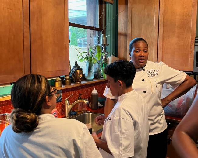 Three individuals in chef uniforms are gathered around a sink in a kitchen, engaged in conversation while preparing food.