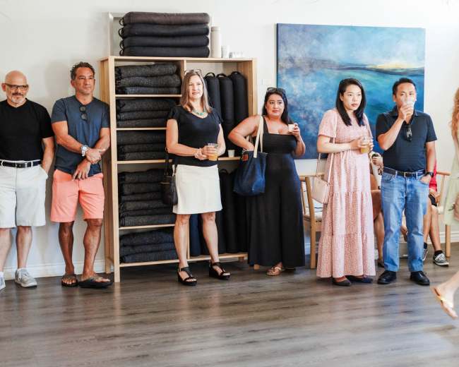 A group of people stands in a well-lit room with a shelf of folded blankets and a blue painting on the wall.