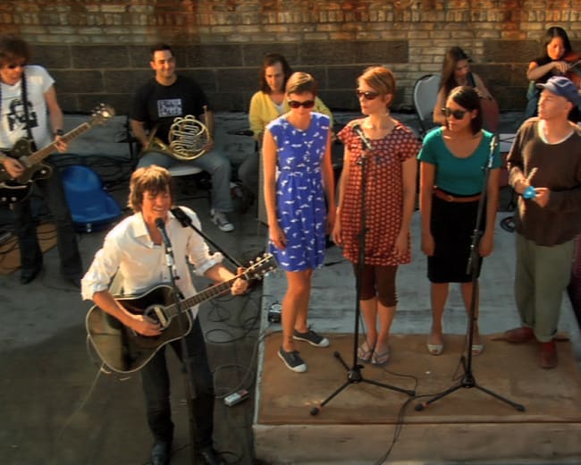 A band performs on an outdoor stage while vocalists sing along, accompanied by musicians playing various instruments.
